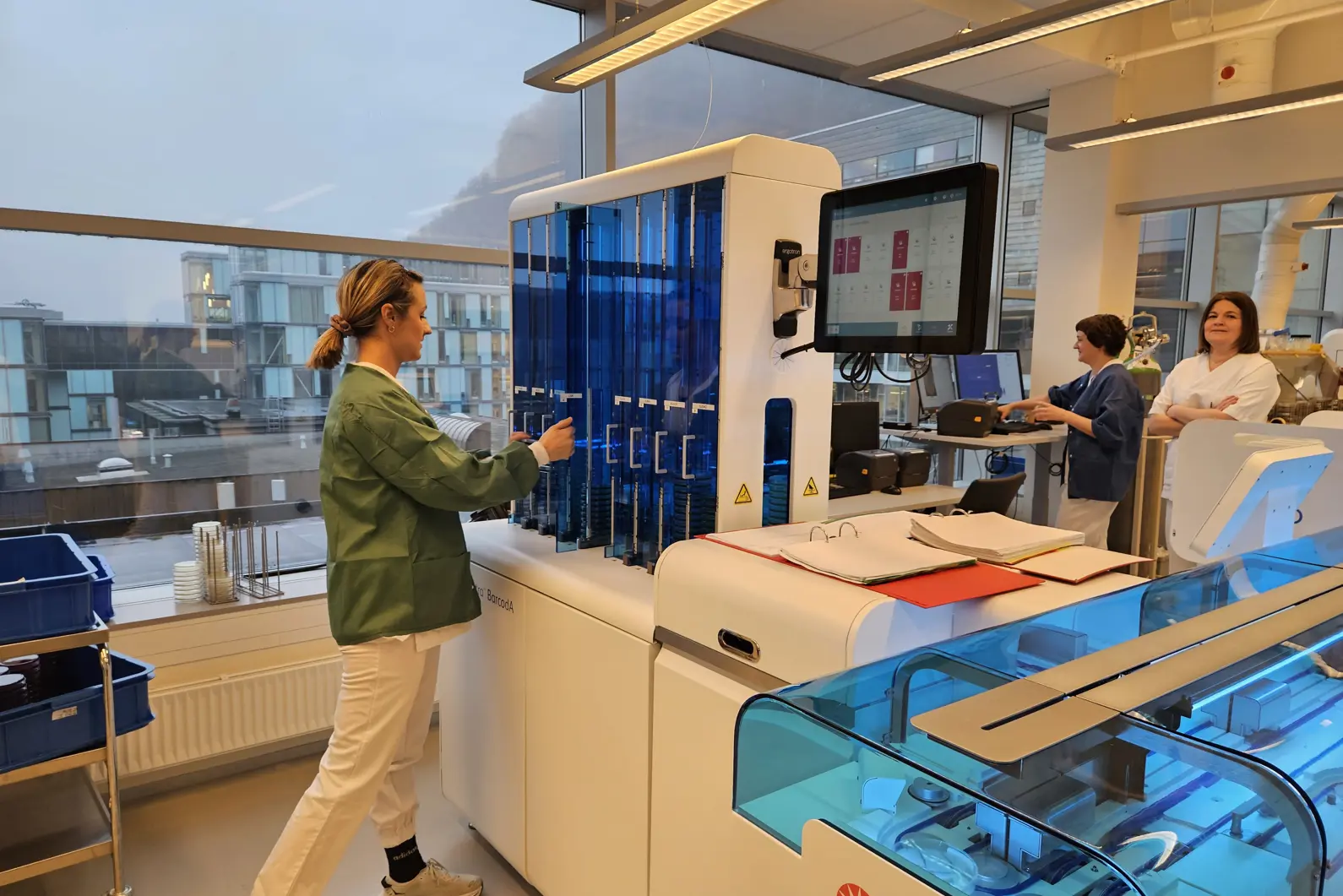 En person i grønn jakke på lab med kollegaer i bakgrunnen