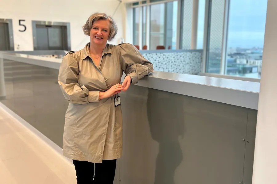 En kvinne står i gangen med grått hår og beige skjorte